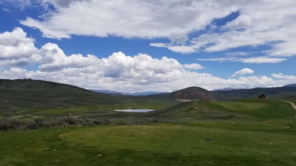 The property sits in a stunning natural landscape with rolling hills, a serene pond, and distant mountain peaks under expansive skies.