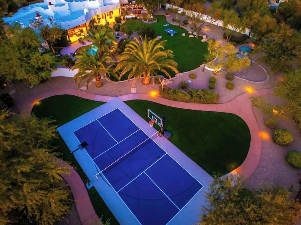 An aerial view reveals the property's impressive grounds at dusk, featuring a full-sized tennis court, lush lawns, mature palm trees, and landscaped pathways with ambient lighting surrounding the main building.