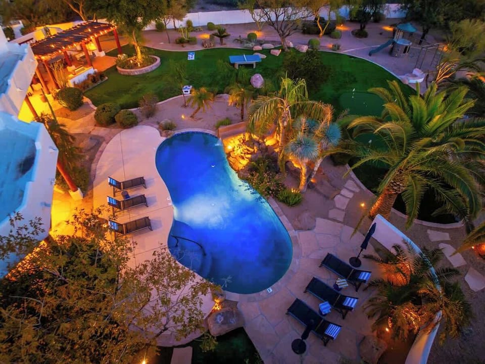 Aerial view of the resort's lush grounds at twilight, showcasing a curved pool, manicured lawns, outdoor seating pavilions, and tropical landscaping illuminated by warm lighting throughout the property.