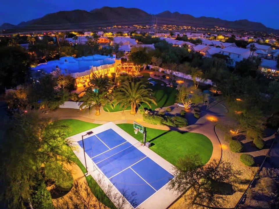 Aerial view at dusk showcases the property's expansive grounds featuring a private tennis court, lush palm trees, resort-style pool, and glowing outdoor spaces nestled within a residential neighborhood backed by dramatic mountain scenery.