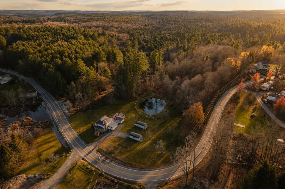 This aerial view shows the property tucked along a quiet country road, surrounded by forest with a private pond just steps from the house and a couple of storage sheds out back for gear.