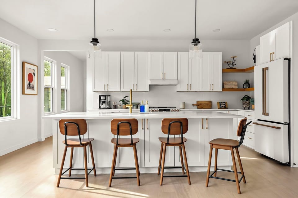 Sleek modern kitchen with white cabinetry, gold hardware, a spacious island with a farmhouse sink, pendant lighting, four wooden barstools, a stainless steel gas stove, a built-in refrigerator, open wooden shelves with decor, and a coffee station.
