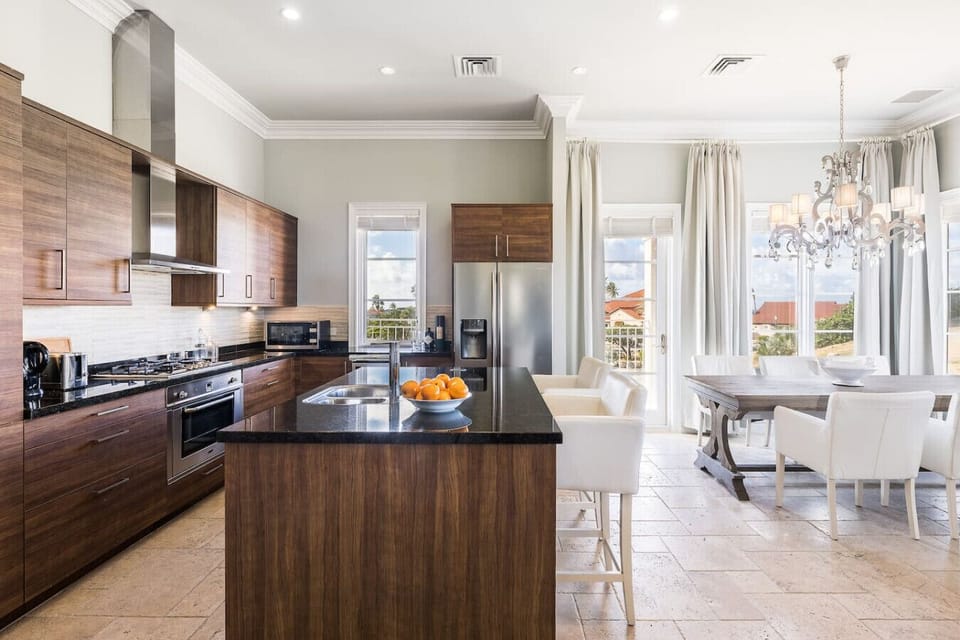 A sleek kitchen with wood cabinetry, black granite counters, and a spacious island flows seamlessly into a bright dining area featuring a rustic table, white chairs, and a chandelier. Large windows fill the space with natural light and scenic views.