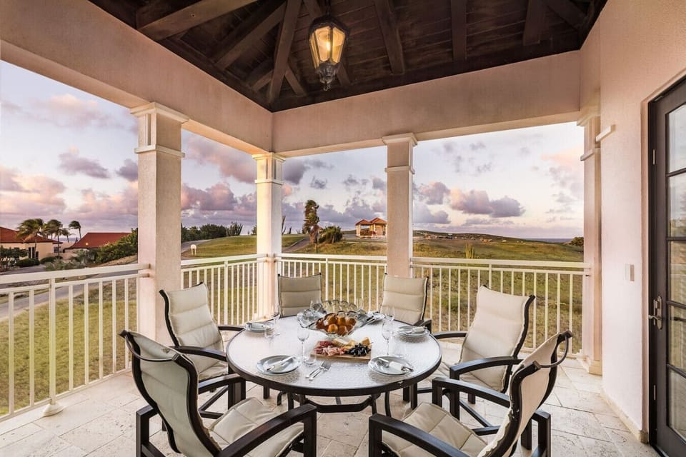 A charming outdoor dining area with a round table and seating for six, offering stunning sunset views and surrounded by elegant columns under a vaulted ceiling.