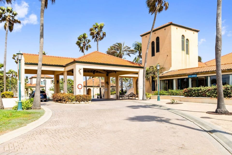 The entrance features a Mediterranean-inspired design with terracotta roofs, palm trees, and an inviting open-air structure, creating a warm and welcoming atmosphere.