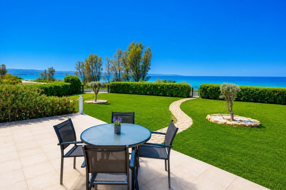 A ground-level patio perspective of the beachfront property, highlighting a comfortable outdoor dining setup and the lush lawn layout leading toward the coast.