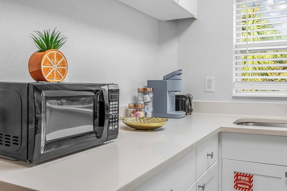 Compact kitchen counter with microwave, coffee maker, and natural light from window blinds.