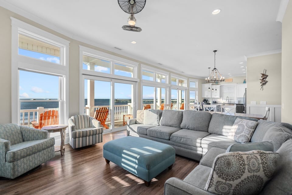 Bright oceanfront living room in Oak Island, NC with gray sectional, blue ottoman, patterned chairs, hardwood floors, and wall of windows.
