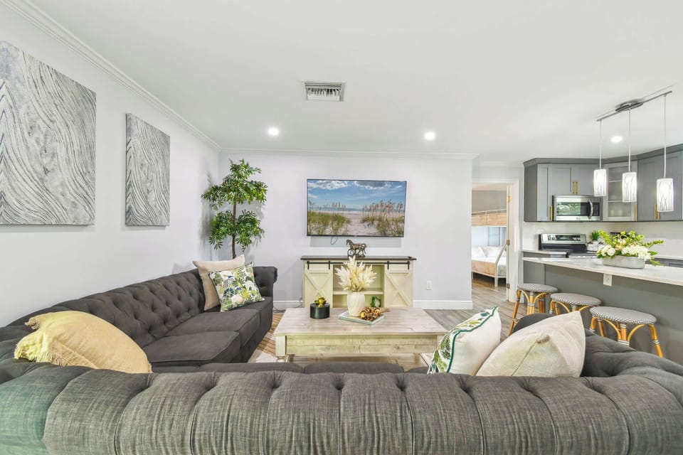 Elegant Largo pool home near Indian rocks beach living room showcasing plush tufted sectional, coastal artwork, and modern farmhouse décor. Open-concept design with kitchen access and premium entertainment setup. 