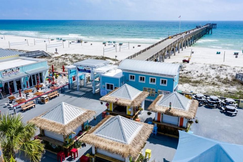 Beachfront paradise with colorful buildings, fishing pier, and pristine white sand stretching along turquoise waters.