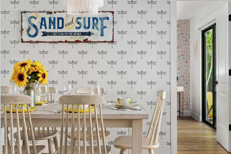 This close-up captures the same dining area, focusing on the stylish "Sand and Surf" sign adorning the wall. Sunflowers on the table create a warm atmosphere, while the dragonfly-patterned wallpaper adds a playful touch.