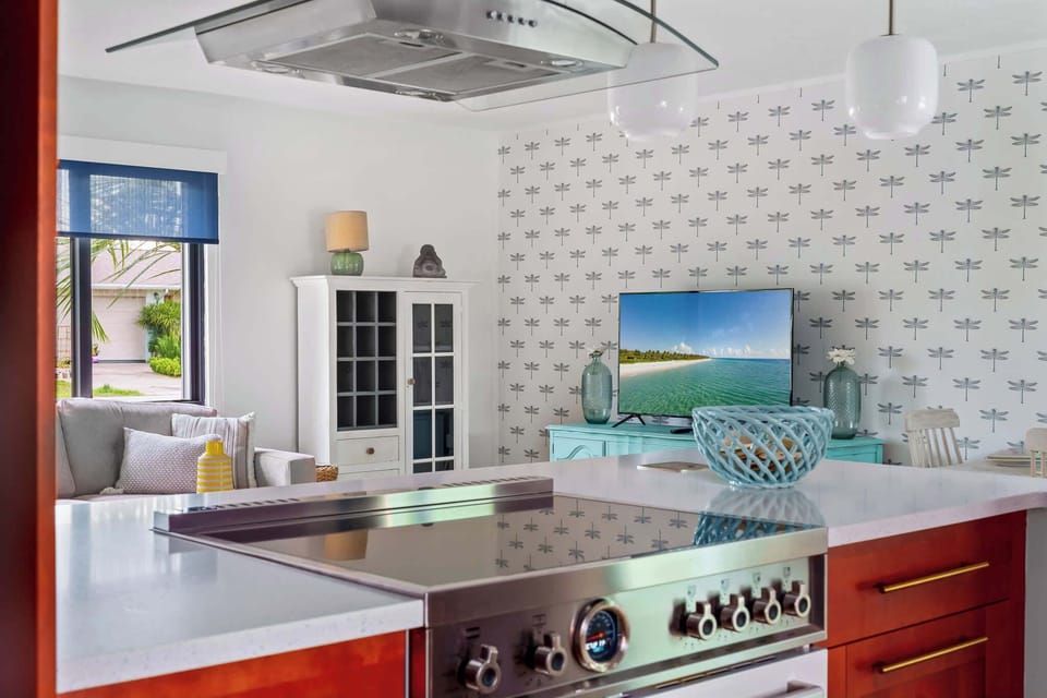  This photo offers a glimpse of the kitchen from a different angle, highlighting a stainless steel gas range and a mounted hood. A vibrant television displays a serene beach view, adding to the light, airy feel. 