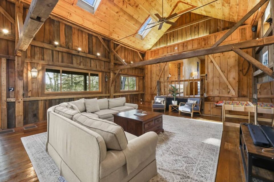 The living area features a large sectional sofa, flat-screen TV, foosball table, and cozy lounge chairs in an open, wood-beamed space.