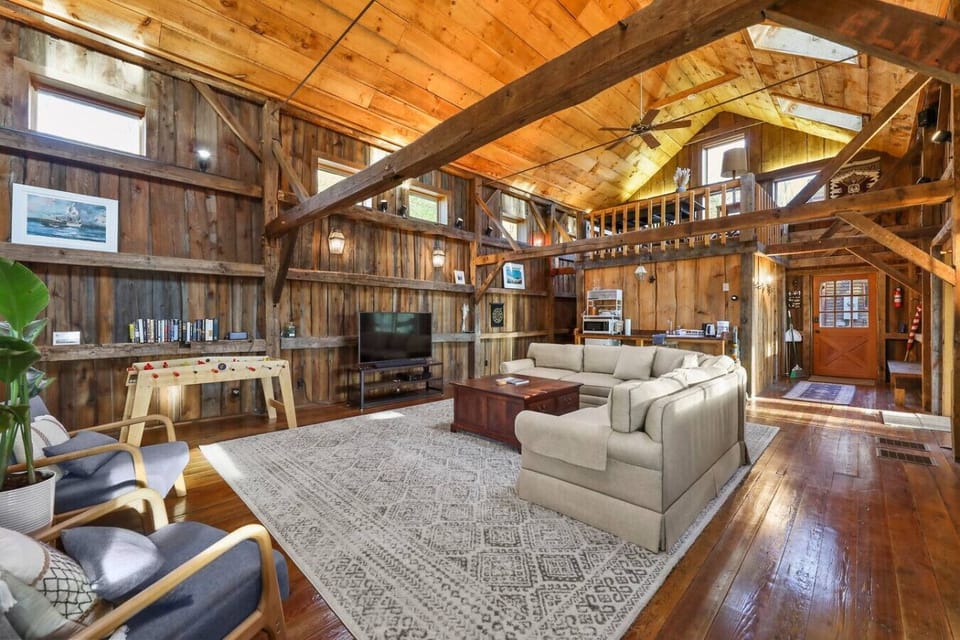 The living area features a large sectional sofa, flat-screen TV, foosball table, and cozy lounge chairs in an open, wood-beamed space.