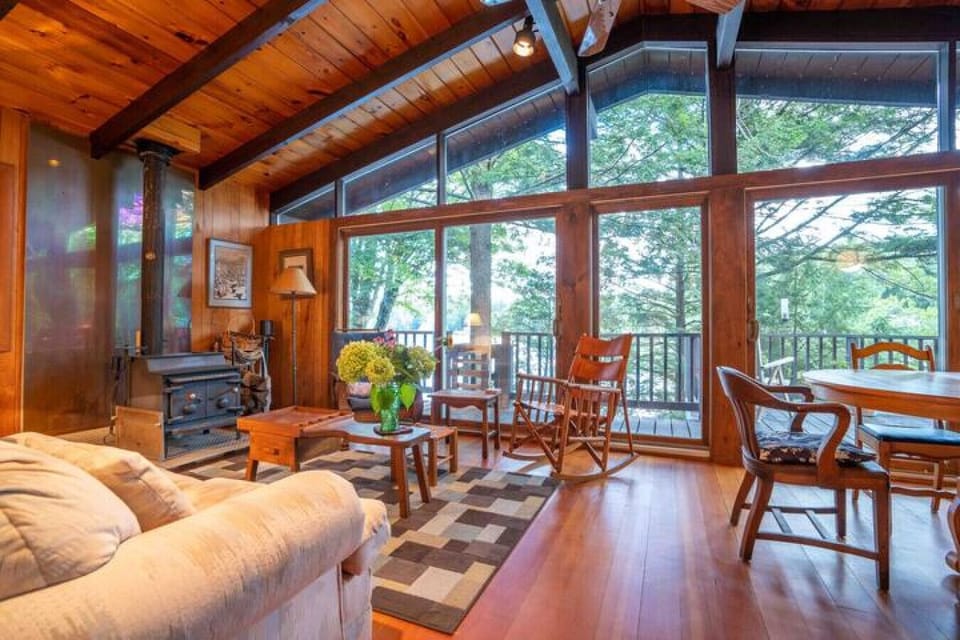 A bright, open living room with vaulted ceilings, a wood-burning stove, and floor-to-ceiling windows overlooking the trees.