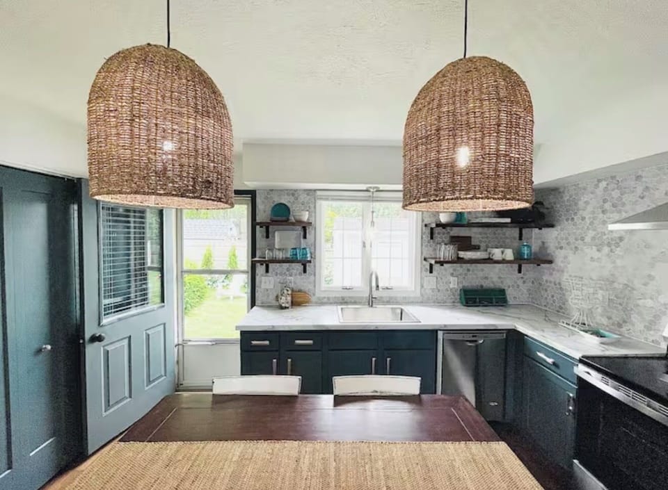 This elegant kitchen corner showcases natural light streaming through large windows, beautifully illuminating the sleek teal cabinetry and open shelving. Woven pendant lights add a warm, rustic charm to the modern design.