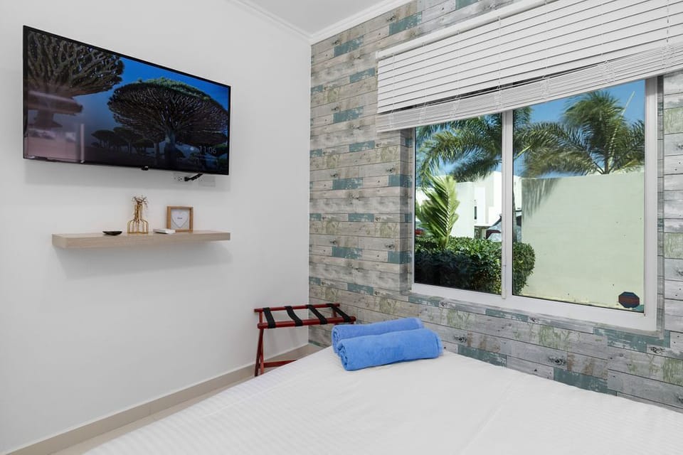 Your serene bedroom retreat offers modern comfort with a wall-mounted TV, climate control, and a bright window framing tropical palms.