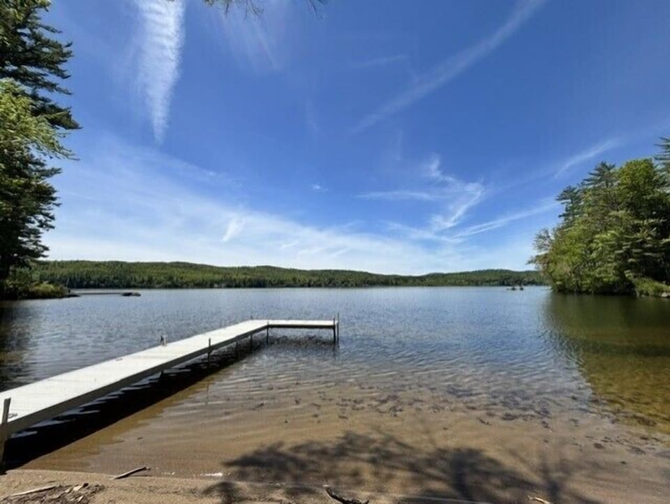 A tranquil lakefront setting featuring a private dock extending into clear waters, surrounded by lush forested shores under expansive summer skies.