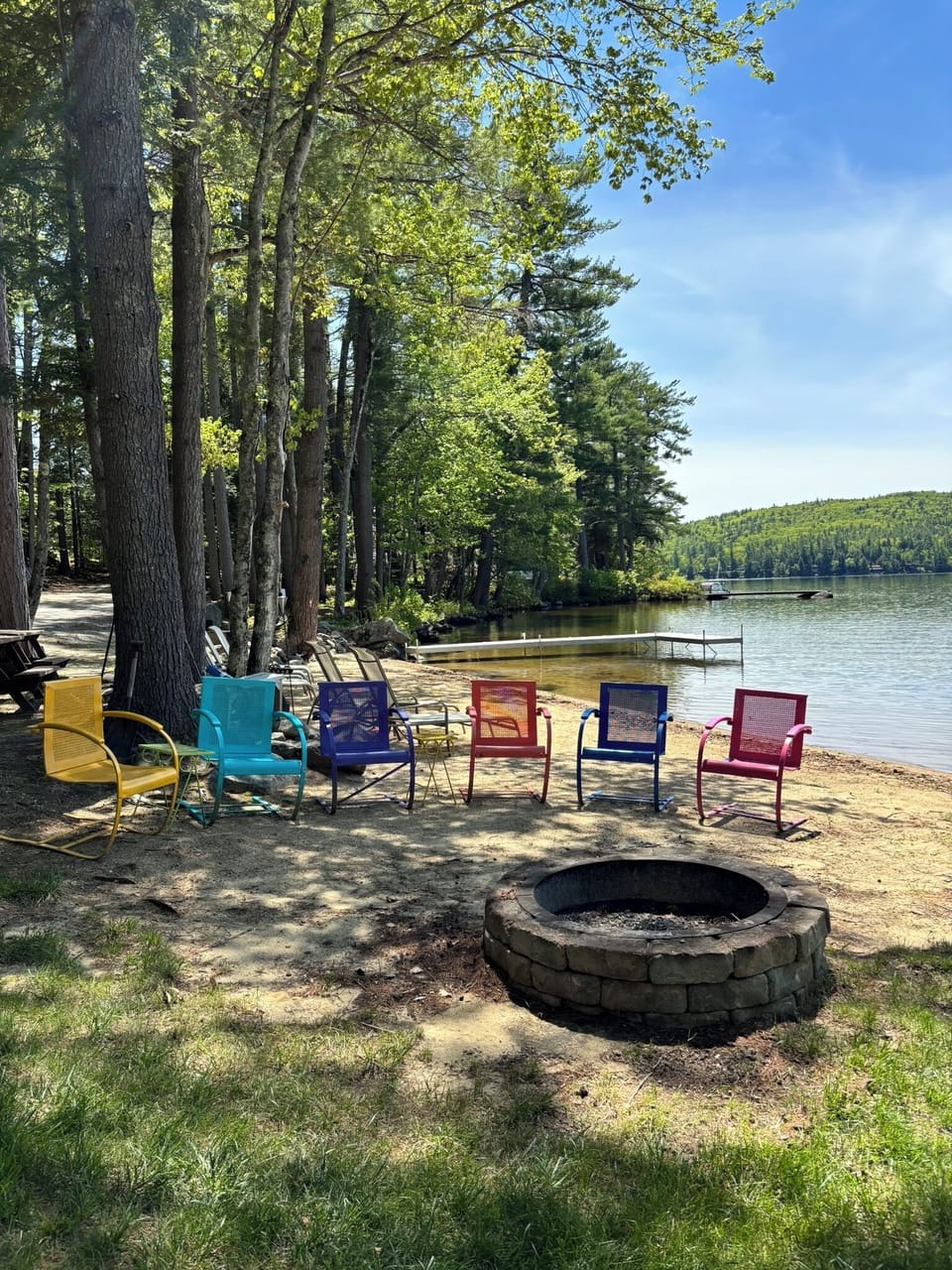 Gather by your private lakefront fire pit and unwind in colorful chairs, where tranquil waters and a gentle breeze under tall trees await your evenings.