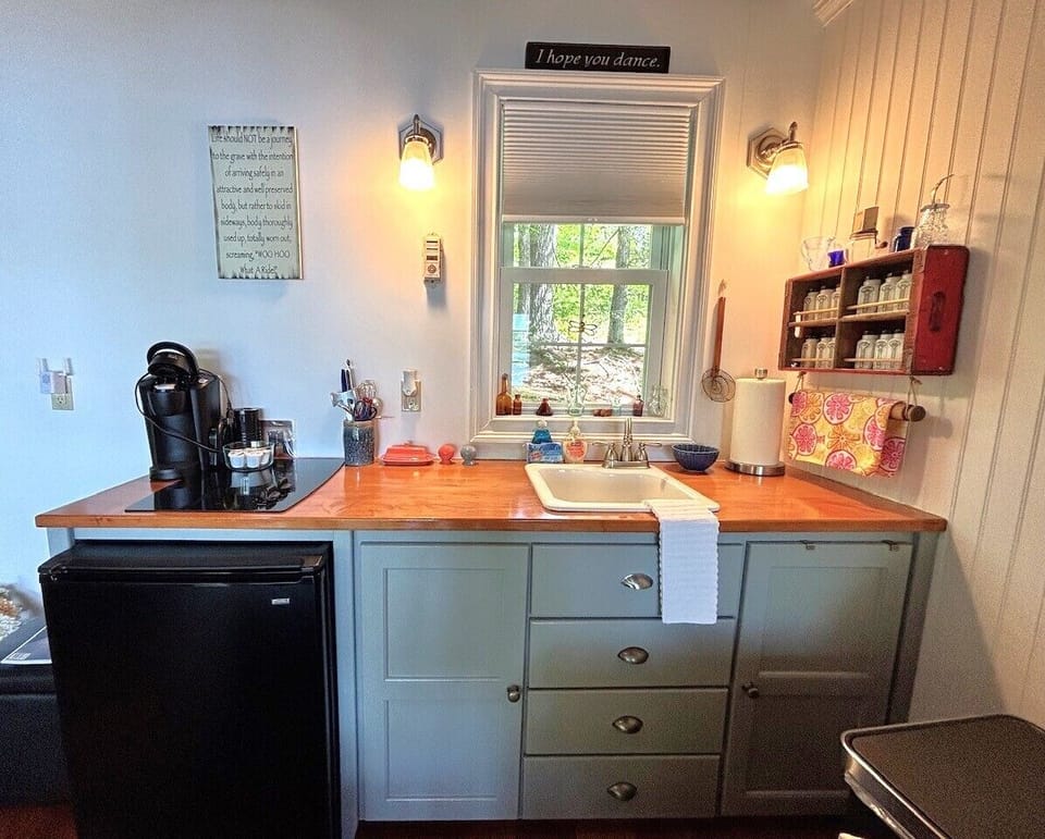 Your cozy kitchenette features warm wood countertops, a mini-fridge, coffee maker, and a charming window overlooking greenery—perfect for preparing morning coffee or light snacks during your stay.