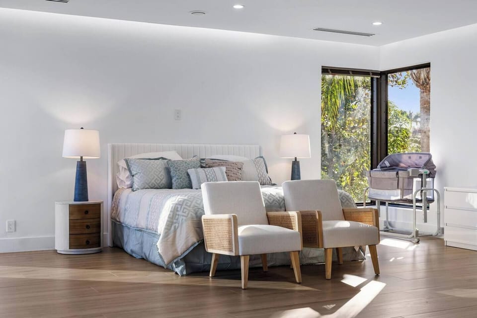 Sunlit primary suite with upholstered platform bed, rattan accent chairs, and corner glass doors opening to tropical greenery