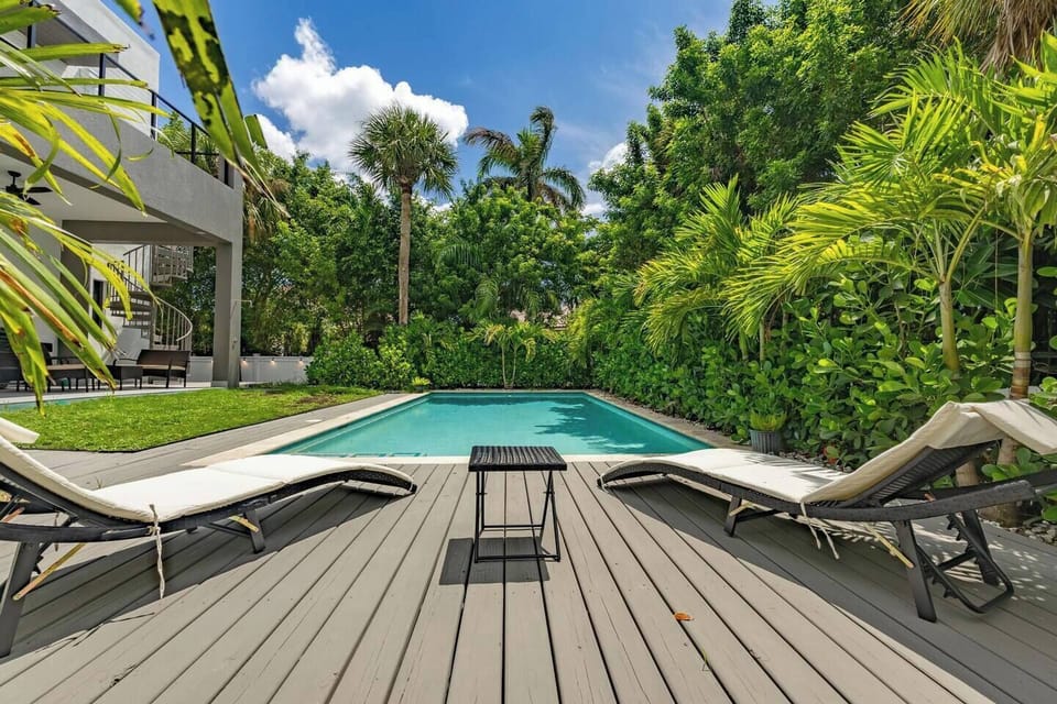 Sunny heated pool with wood deck, pair of lounge chairs, and lush tropical privacy hedge — your own backyard oasis