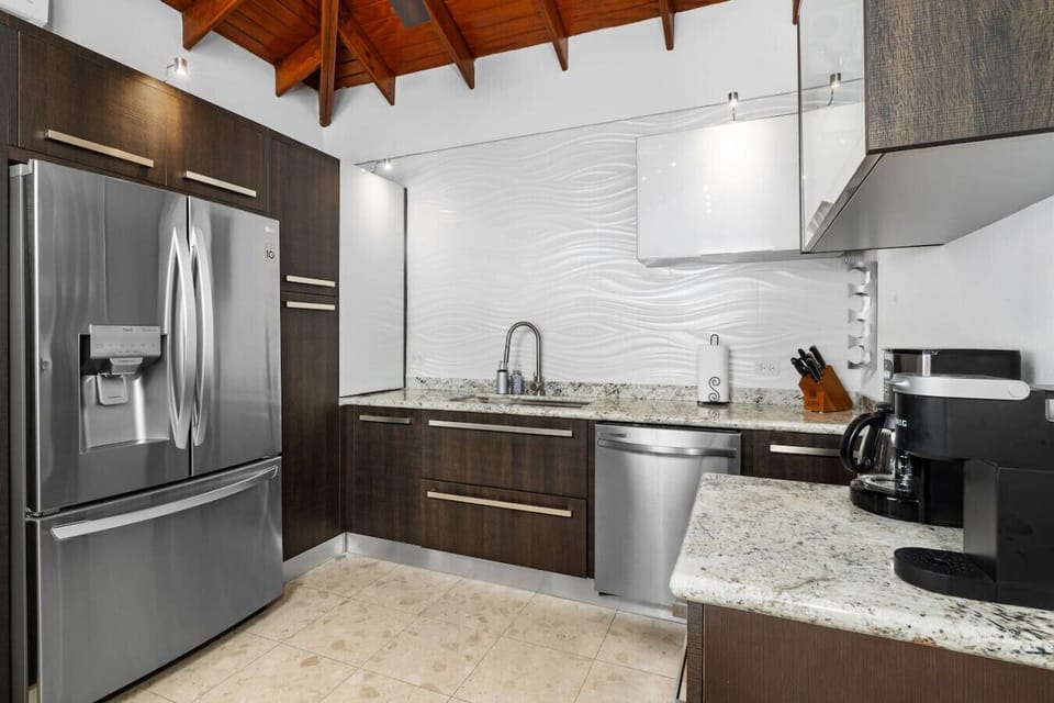 A contemporary kitchen featuring sleek dark wood cabinetry, stainless steel appliances, and granite countertops. The space is complemented by a textured white backsplash and a vaulted wooden ceiling, blending modernity with warmth.
