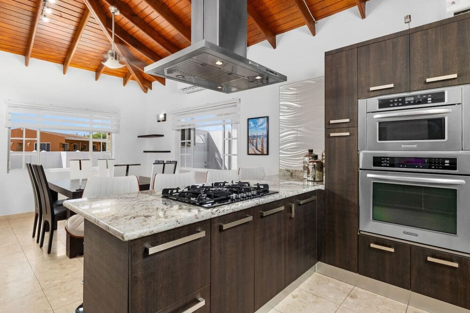 A modern kitchen with a wooden vaulted ceiling, a marble island with a gas stove, built-in stainless steel ovens, and an open dining area featuring white chairs and large windows for natural light.