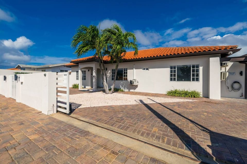 A modern single-story house with white walls and a red-tiled roof, surrounded by a paved driveway and minimal landscaping, featuring palm trees and a clean, minimalist exterior design.
