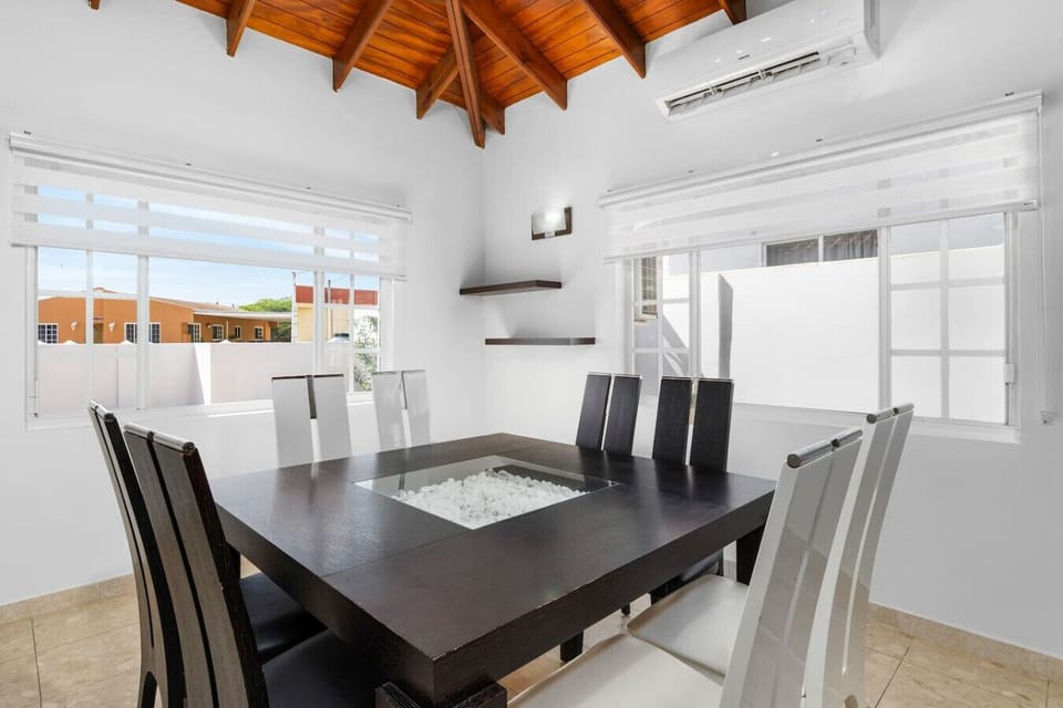 A bright and airy dining room with a modern dark wood table surrounded by sleek white chairs. The space is enhanced by natural light from large windows, a vaulted wooden ceiling, and minimalist decor.