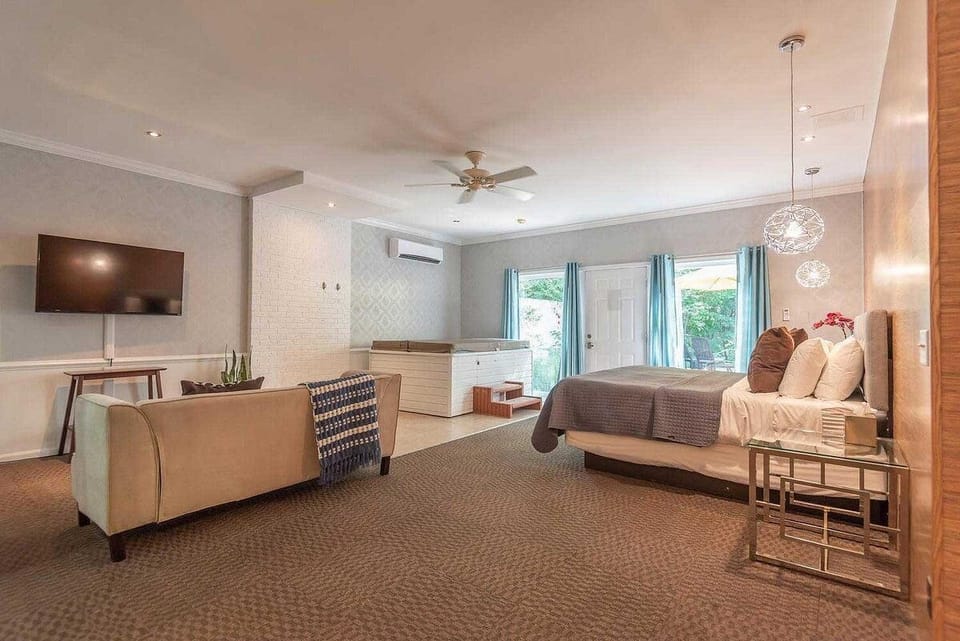 Suite seating area with beige sofa and wall-mounted TV faces a king bed and white brick hot tub near glass doors.