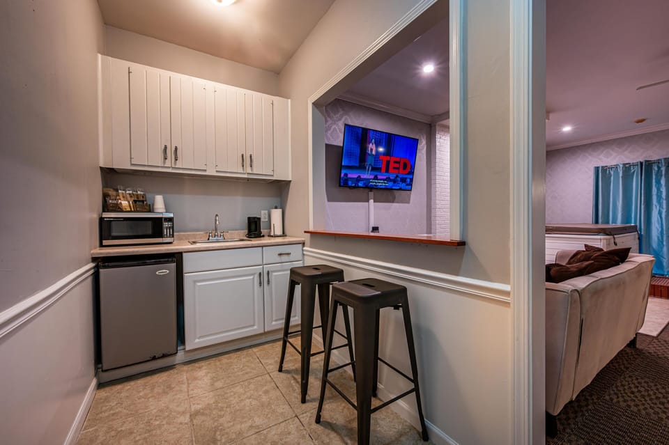 Compact kitchenette with white cabinetry, beige countertop, mini fridge, microwave, and two black metal barstools.