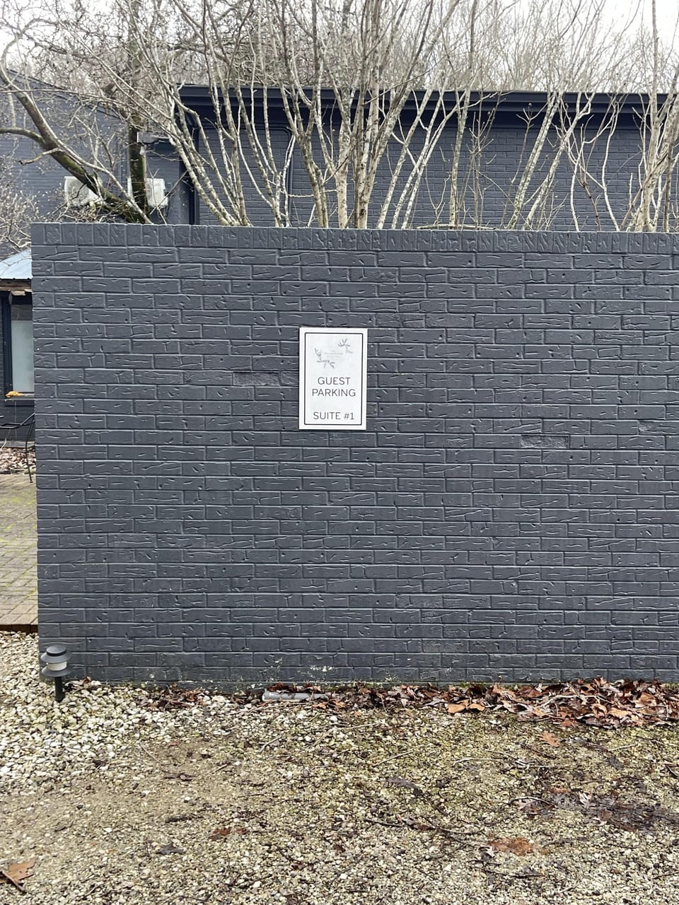 Gray brick wall enclosing guest parking for Suite #1, bordered by gravel and dry leaves under leafless trees.