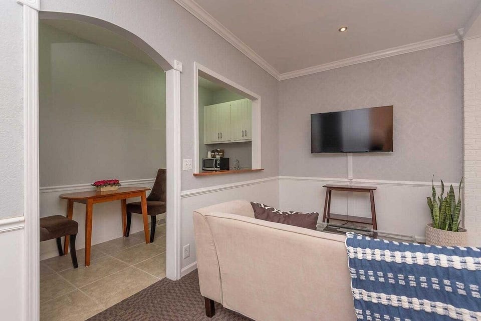 Living area with beige sofa facing a wall-mounted TV, adjacent to a small dining nook with wooden table and brown chairs.
