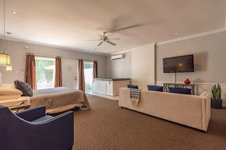 Suite living area with beige sofa, navy armchair, wall-mounted TV, and white hot tub by sliding glass doors.