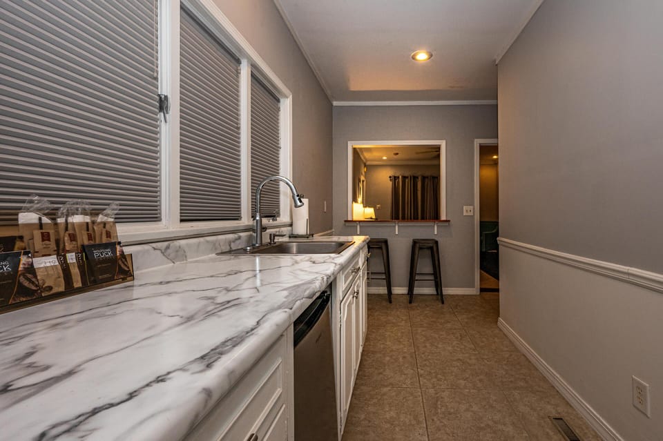 Long marble countertop with stainless steel sink under closed blinds, leading to a breakfast bar with two stools.