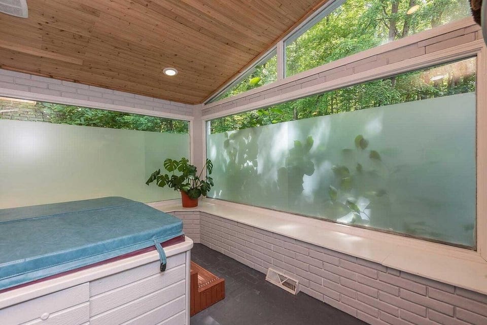 In-room hot tub with white brick walls, frosted windows, vaulted wood ceiling, and a corner plant.