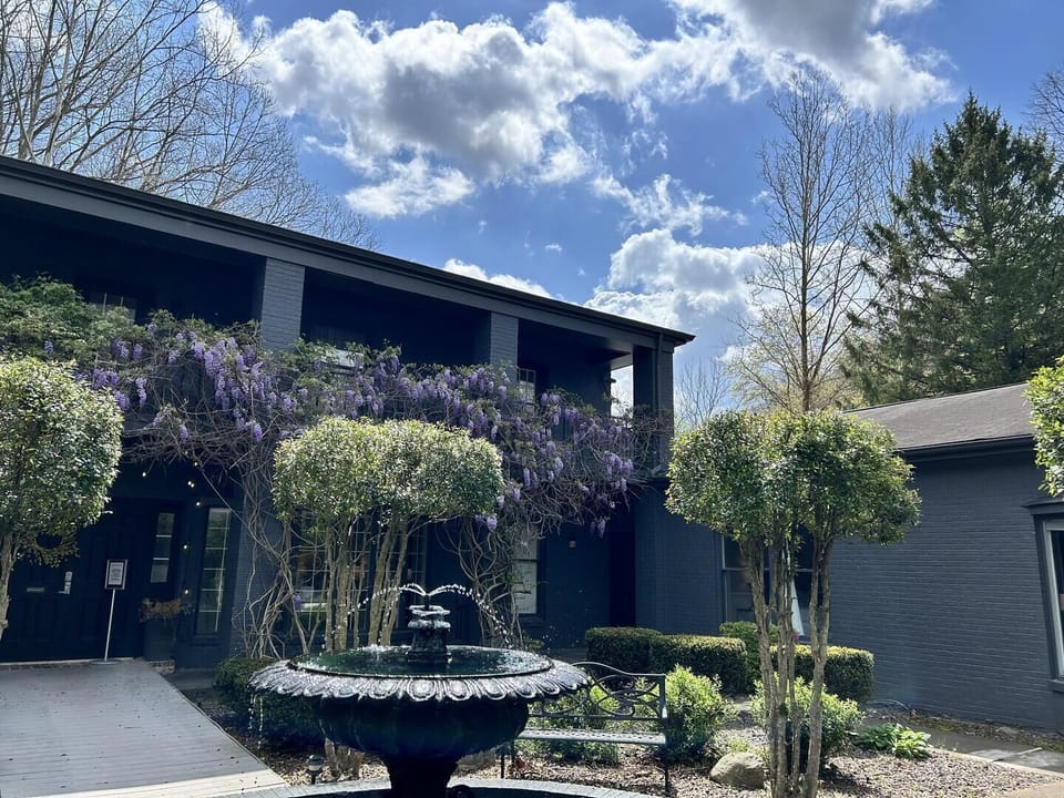 Wisteria vines drape over the dark brick facade with a black metal fountain and trimmed trees in front.