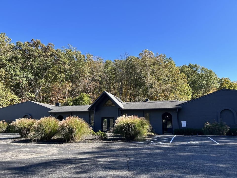 Dark brick inn exterior with triangular glass entry, surrounded by ornamental grasses and mature trees.