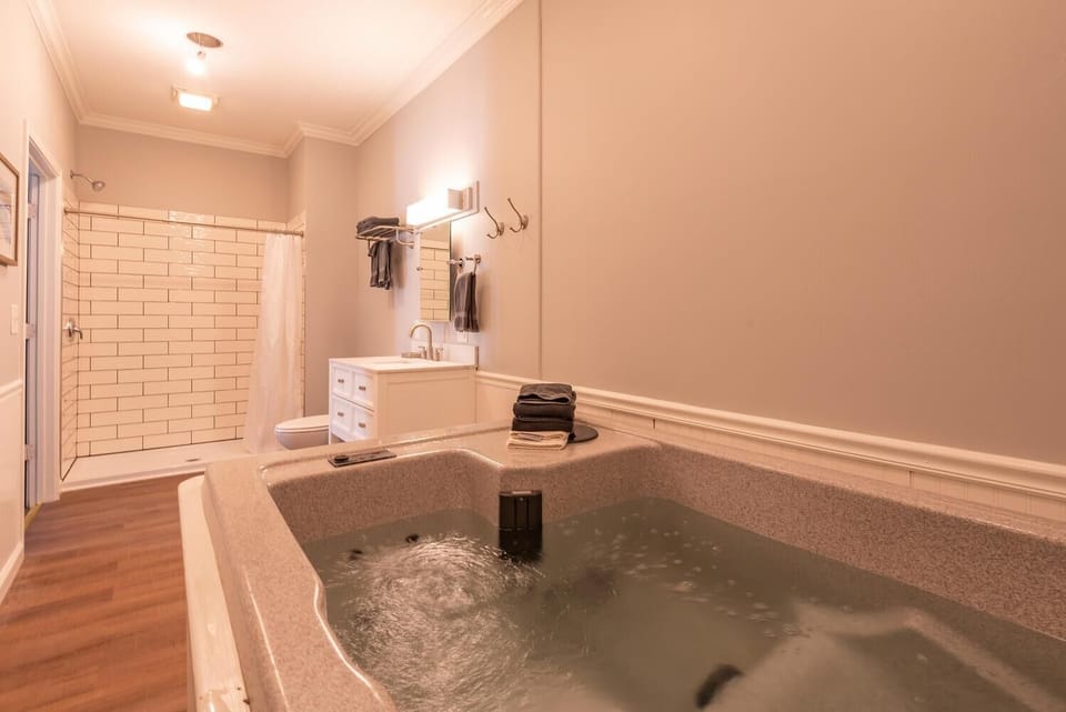 In-room hot tub bubbles beside a white vanity, shower with subway tile, and wood-look flooring in the bathroom.