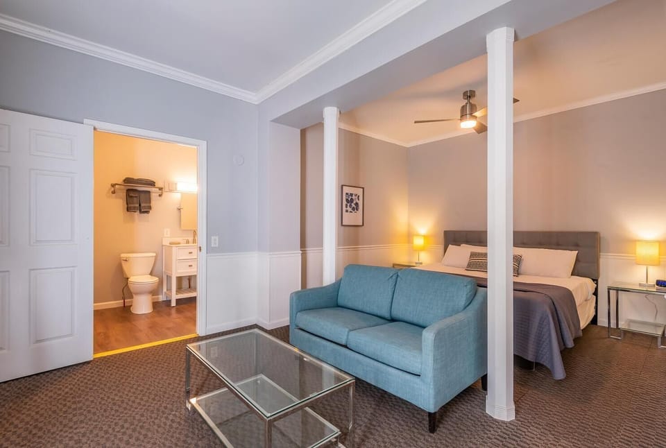 Living area with blue loveseat and glass coffee table, leading into bathroom with white vanity and toilet.