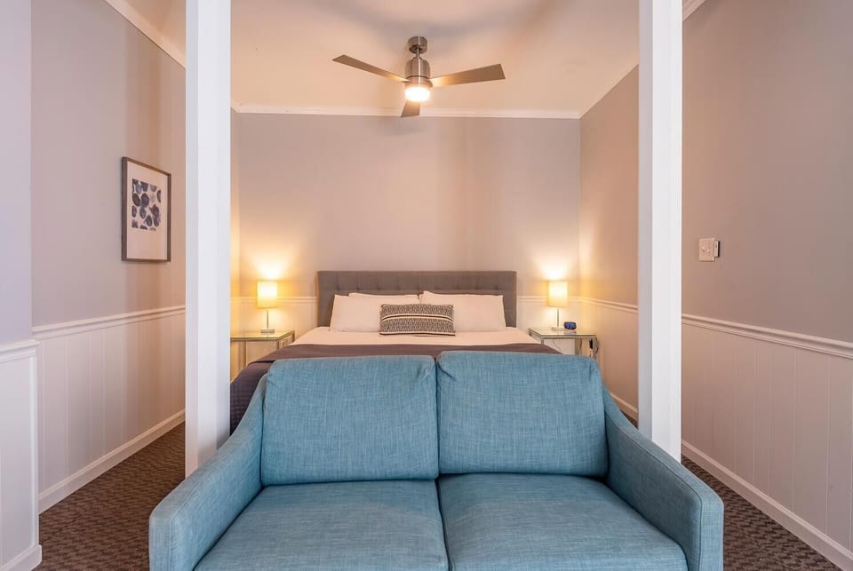 A gray upholstered bed with white bedding sits behind a blue loveseat in the softly lit bedroom.