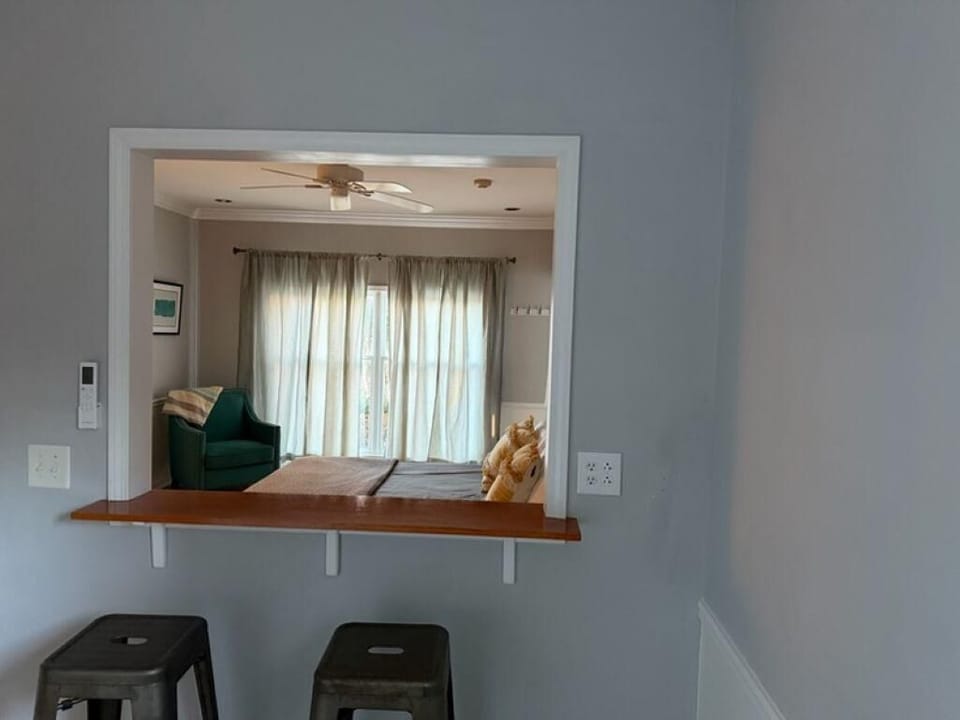 Wood counter bar with metal stools faces an open window into the bedroom with sheer curtains and a teal armchair.