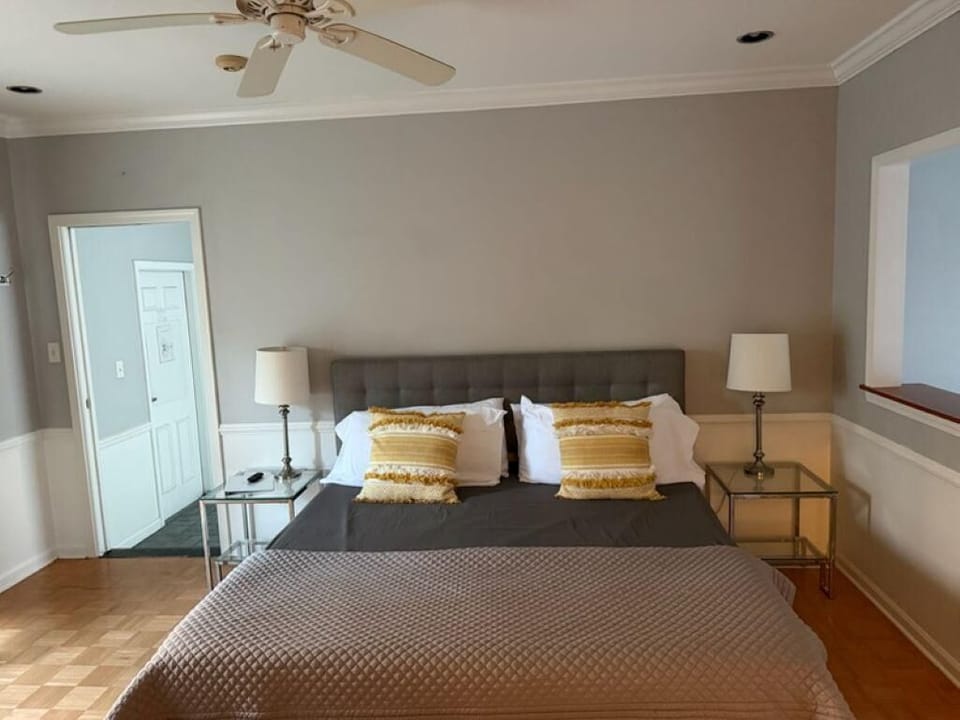 Gray tufted headboard anchors the bed framed by glass nightstands and table lamps with white shades.