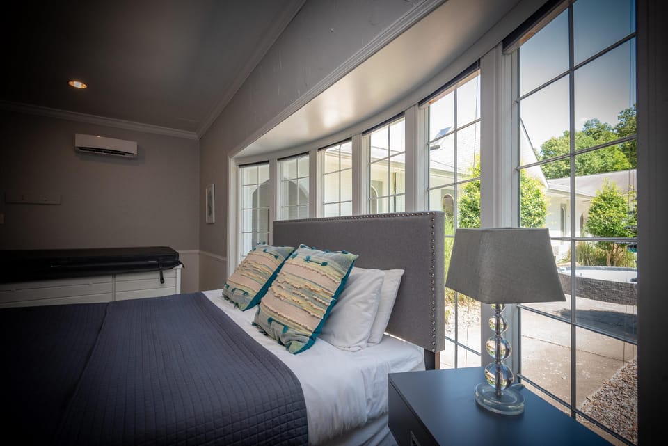 A clear glass lamp with a silver base sits on a black nightstand beside the bed near large grid-pane windows.