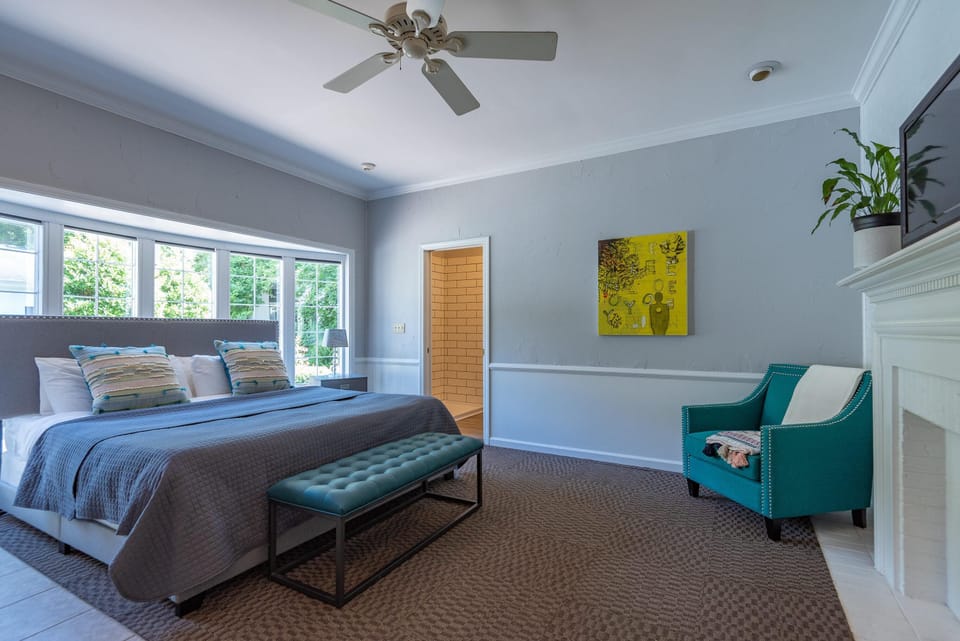 King bed with gray quilted bedding faces a teal tufted bench and large windows in the bedroom.