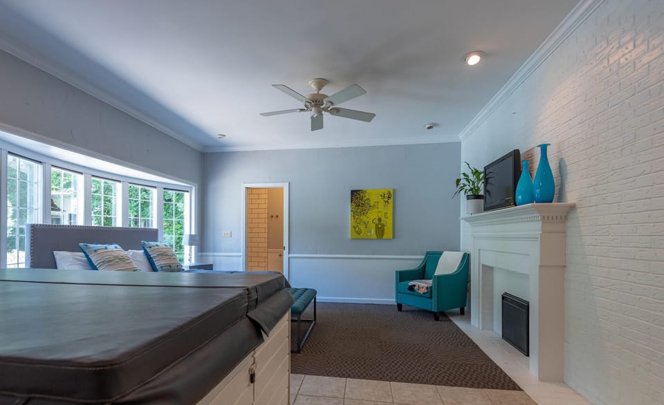 A covered hot tub anchors the foreground with a teal armchair and white brick fireplace in the bedroom area.