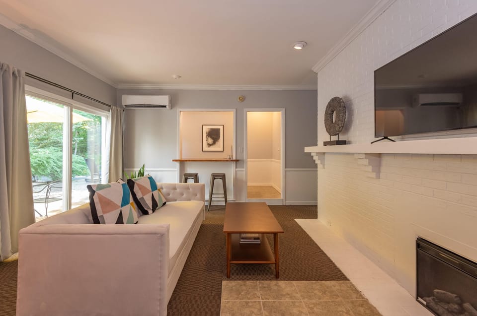 Living area with white tufted sofa, geometric pillows, wood coffee table, and wall-mounted TV over painted brick hearth.
