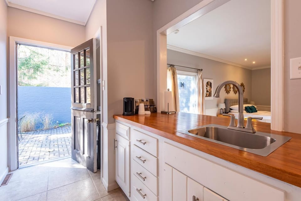 Wood countertop kitchen sink overlooks bedroom with queen bed and beige curtains by sliding door.