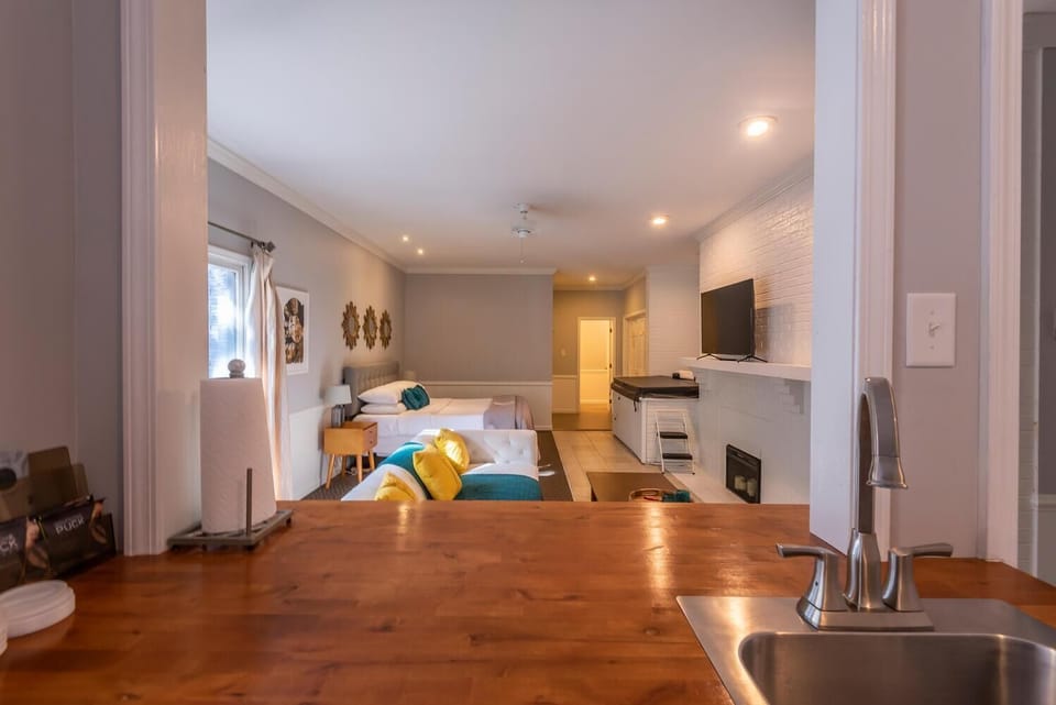 View from kitchen counter over wooden surface and stainless steel sink into suite with white tufted sofa and queen bed.